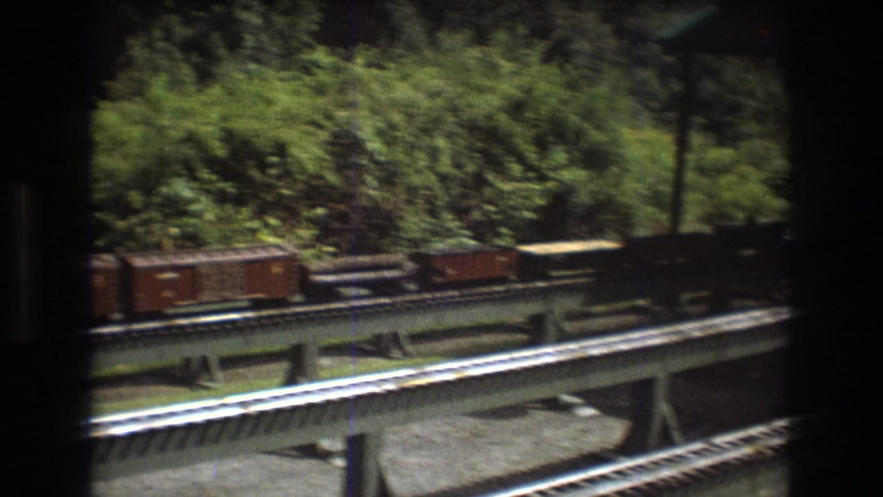 In this Baxter, Maine 1975 8mm frame, brown model freight cars pass across an elevated railroad track with leafy vegetation and dark film borders around the view.
