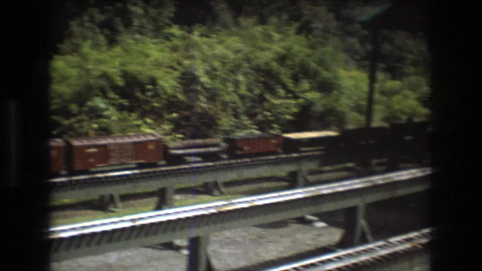 In this Baxter, Maine 1975 8mm frame, brown model freight cars pass across an elevated railroad track with leafy vegetation and dark film borders around the view.