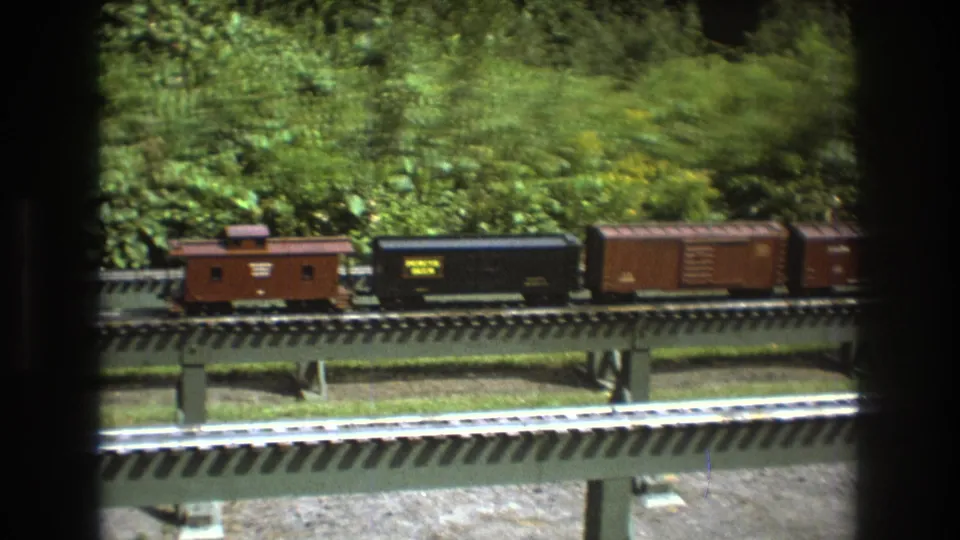 A model train in Baxter, Maine in 1975 shows a caboose, a black lettered freight car, and a brown boxcar crossing an elevated track against dense greenery.
