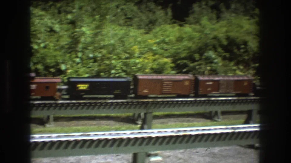 The 1975 Baxter, Maine home-movie frame shows black and brown model freight cars moving past on raised rails, with another track visible in the foreground.