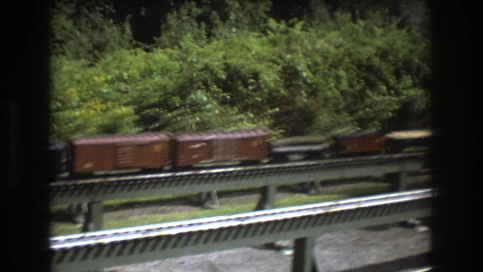 Several brown model freight cars continue across the elevated model railway in this Baxter, Maine 1975 8mm frame, with dense foliage and dark frame edges visible.