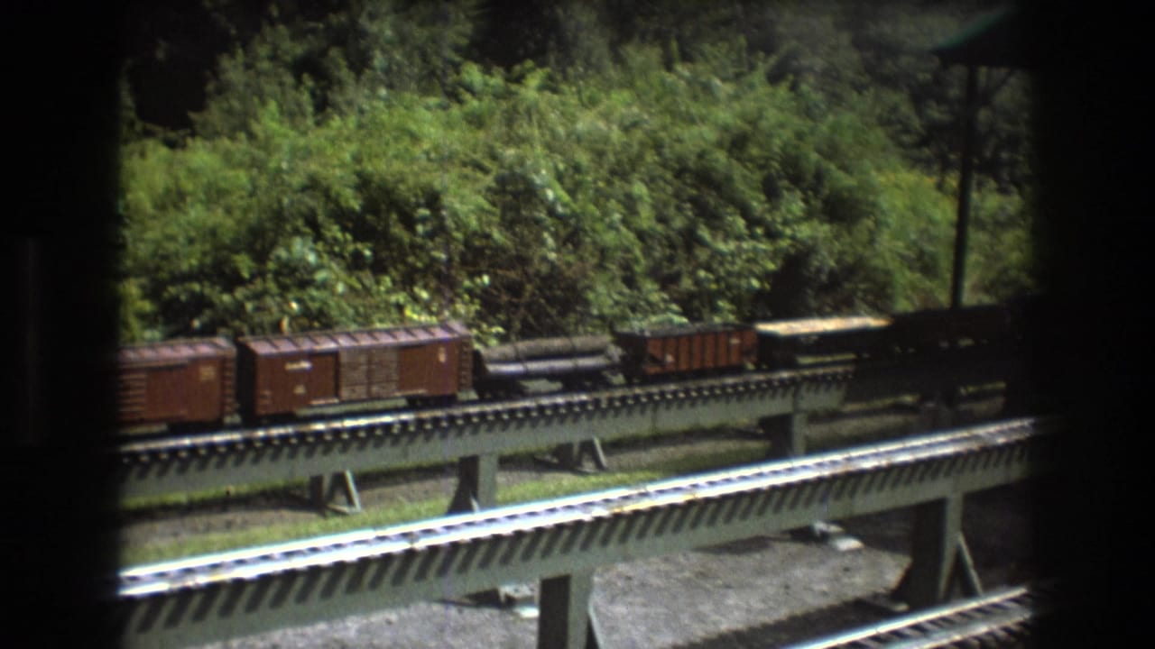 1975 Baxter, Maine home-movie frame showing miniature freight cars and a caboose on elevated outdoor model railroad tracks near dense green foliage.