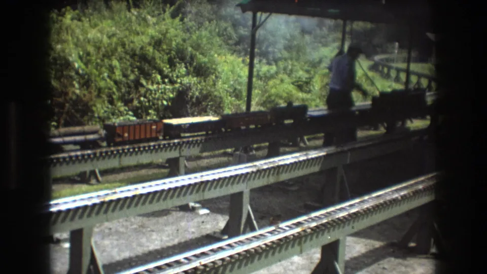 1975 Baxter, Maine home-movie frame showing a man in a light shirt working beside a miniature train under a small shelter.