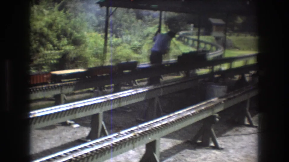 1975 Baxter, Maine frame showing a man beside an outdoor miniature railway with freight cars, elevated rails, and surrounding foliage.