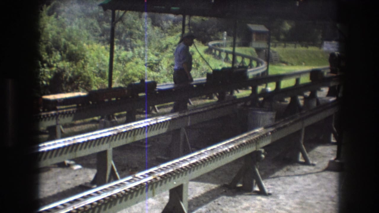 In Baxter, Maine in 1975, a man stands on a miniature train platform beside small train equipment on elevated rails beneath a station roof.