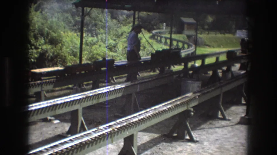 In Baxter, Maine in 1975, a man stands on a miniature train platform beside small train equipment on elevated rails beneath a station roof.