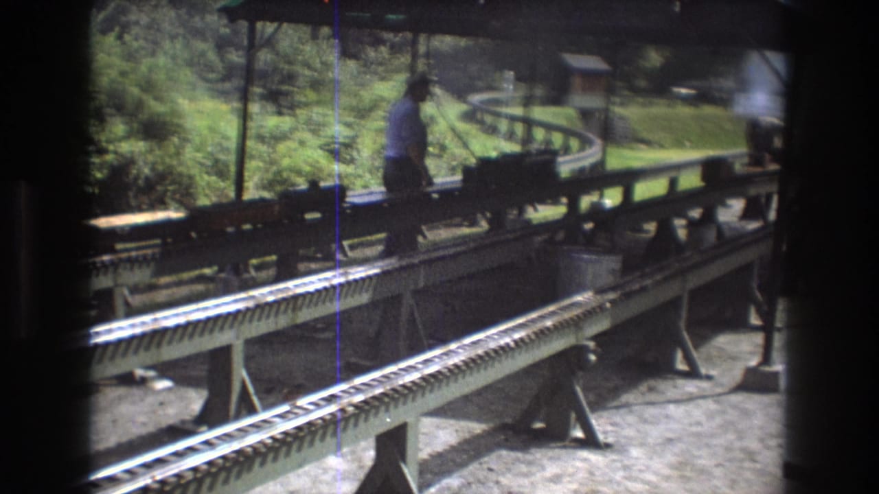 A 1975 home-movie frame shows a man near miniature train equipment on elevated tracks at an outdoor amusement park station in Baxter, Maine.