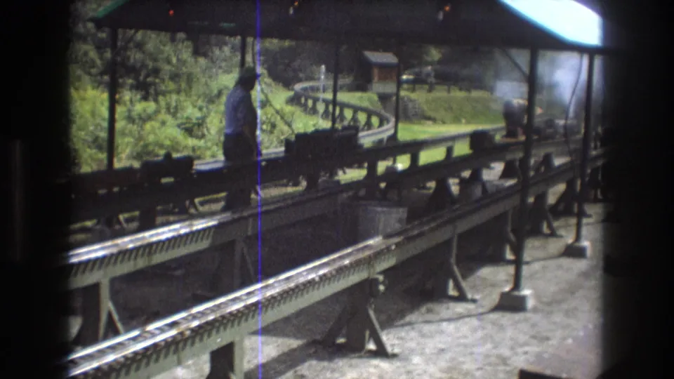 The frame shows the covered station area, foreground rails, and a man standing beside a small train attraction in Baxter, Maine in 1975.
