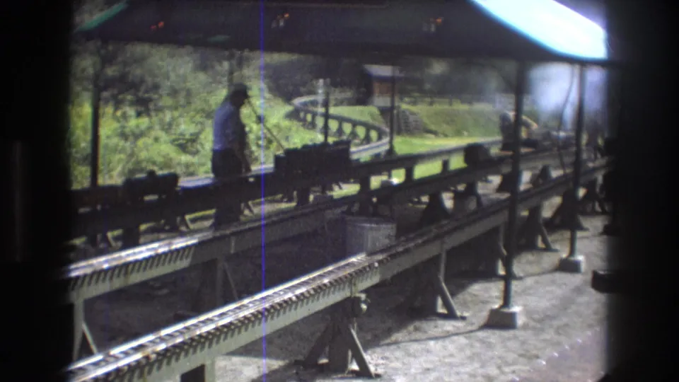 At the end of the clip, the man remains near the miniature train equipment under the roofed platform, with elevated rails and a curve of track visible behind him.