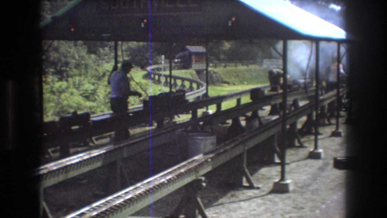 In Baxter, Maine in 1975, an adult worker stands near a small steam train display on elevated wooden tracks beneath a covered platform.