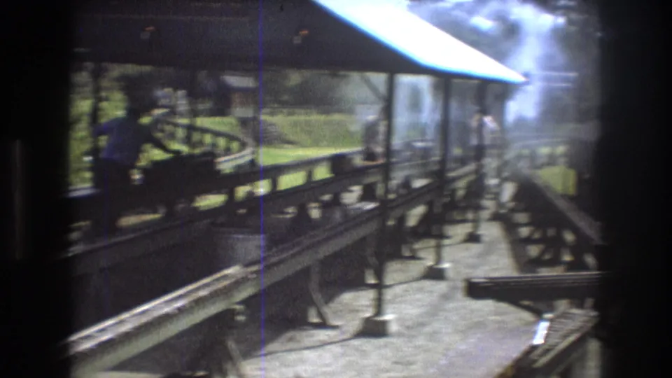 Steam or smoke drifts through the covered miniature train display area in Baxter, Maine, with wooden tracks, posts, and grass visible in the 8mm frame.