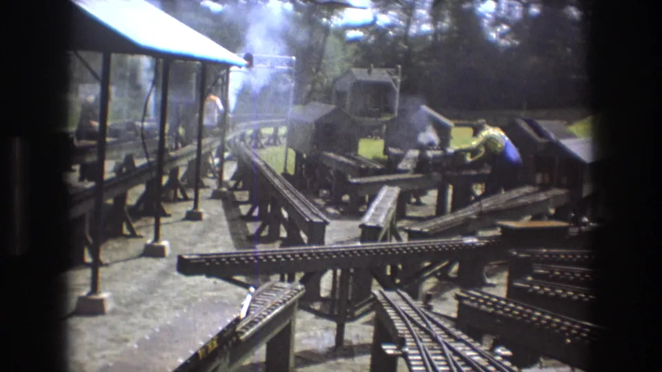 A wider 1975 Baxter, Maine view shows the outdoor miniature steam train layout with intersecting elevated tracks, smoke, a covered platform, and a worker handling equipment.