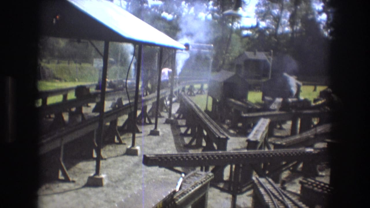 1975 Baxter, Maine 8mm frame showing an outdoor miniature railway layout with tracks, wooden platforms, a small roofed structure, and white steam or smoke rising near the engine.