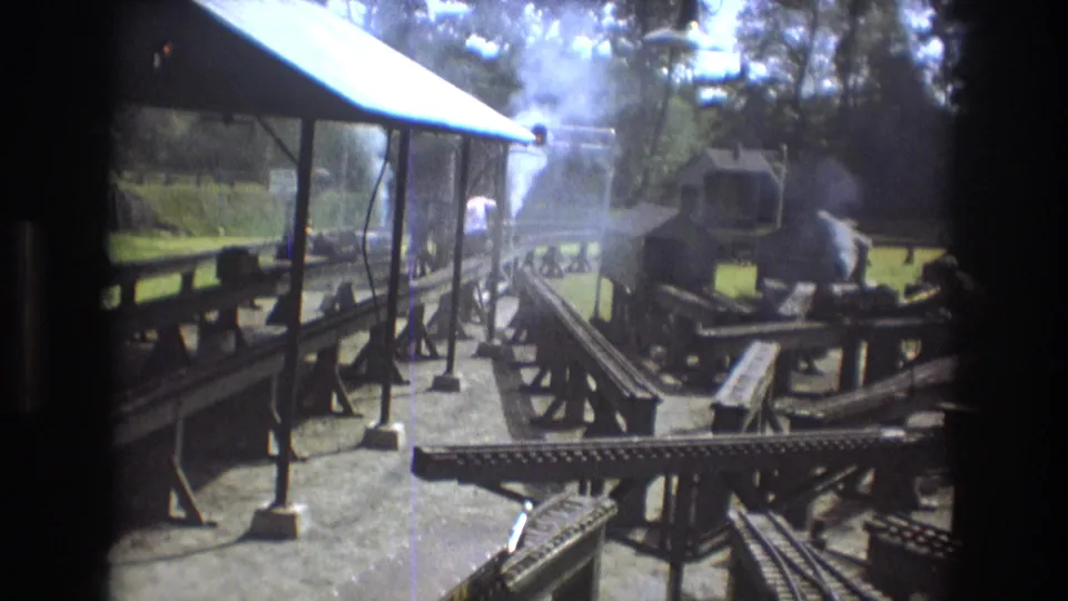 1975 Baxter, Maine 8mm frame showing an outdoor miniature railway layout with tracks, wooden platforms, a small roofed structure, and white steam or smoke rising near the engine.