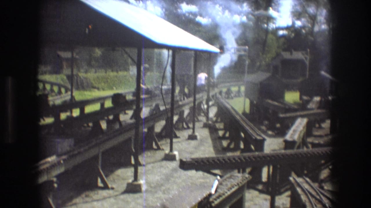 1975 Baxter, Maine 8mm frame of a miniature rail setup with multiple tracks, a small steam engine area, white smoke, a roofed platform, and trees in the background.
