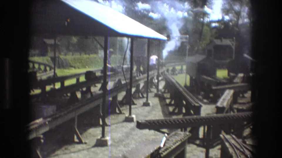 1975 Baxter, Maine 8mm frame of a miniature rail setup with multiple tracks, a small steam engine area, white smoke, a roofed platform, and trees in the background.
