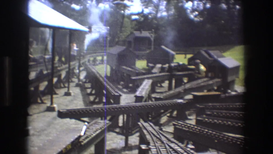 1975 Baxter, Maine 8mm frame showing curving miniature tracks, small shed-like model structures, wooden platforms, and steam drifting above the outdoor railway layout.