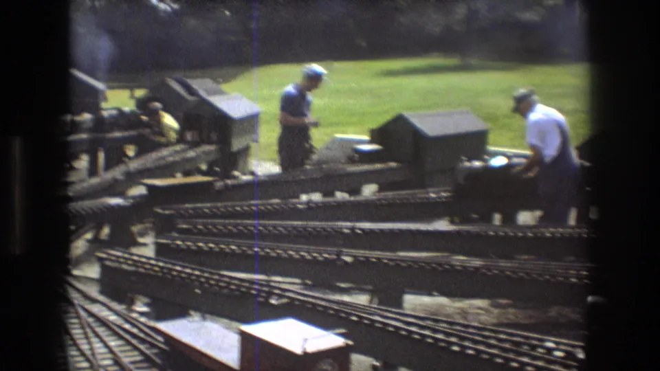 1975 Baxter, Maine 8mm frame showing two adult men near an outdoor miniature railway, with multiple tracks, small buildings, green lawn, and workshop-like rail equipment visible.