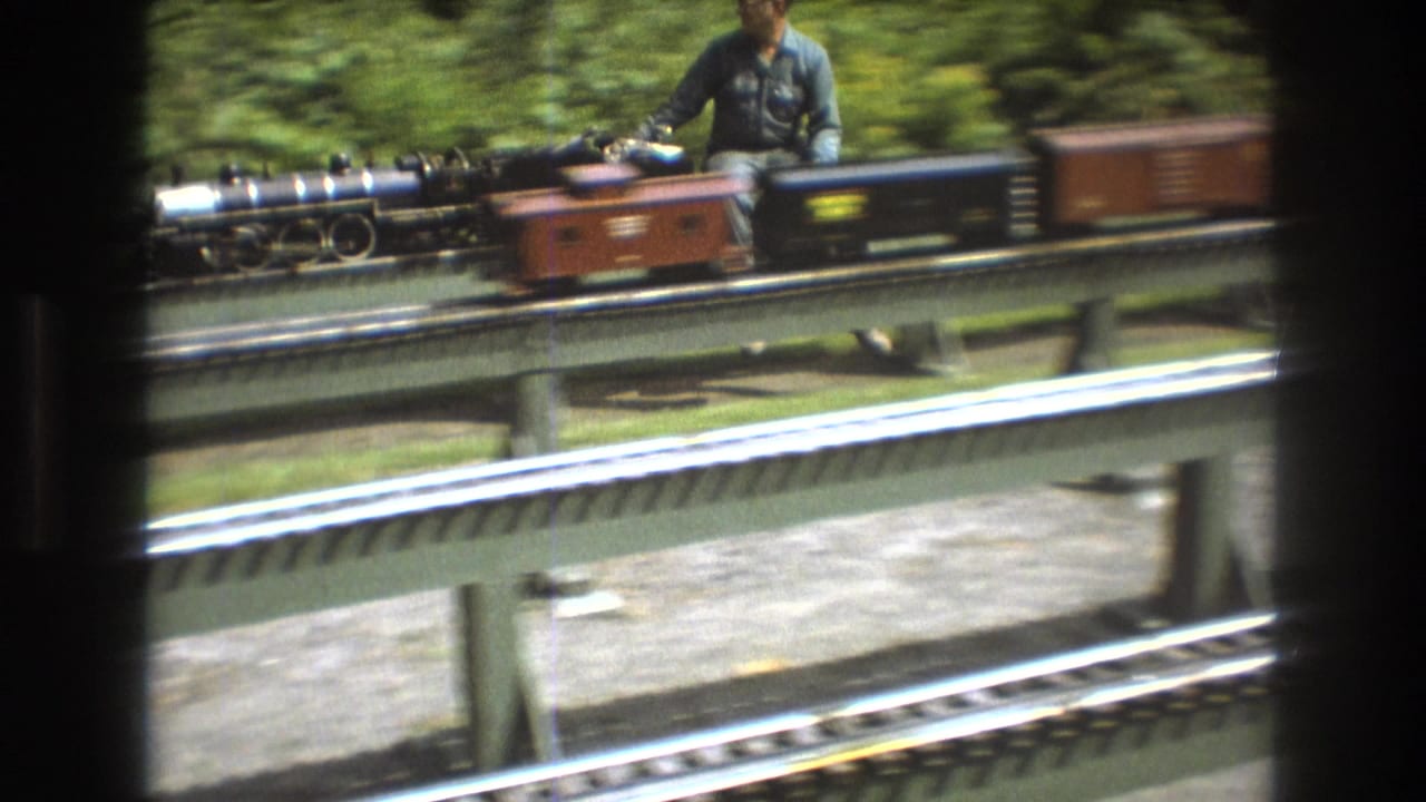In the supplied 1975 Baxter, Maine home-movie context, a miniature steam locomotive with small cars moves along raised green rails as an adult operator is partially visible behind it.