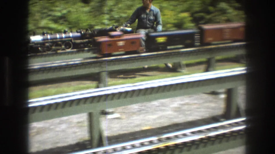 In the supplied 1975 Baxter, Maine home-movie context, a miniature steam locomotive with small cars moves along raised green rails as an adult operator is partially visible behind it.