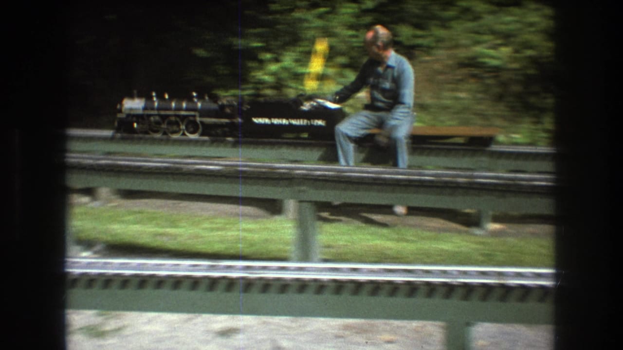 Adult man rides beside a black miniature steam locomotive on an elevated track in a green outdoor setting.