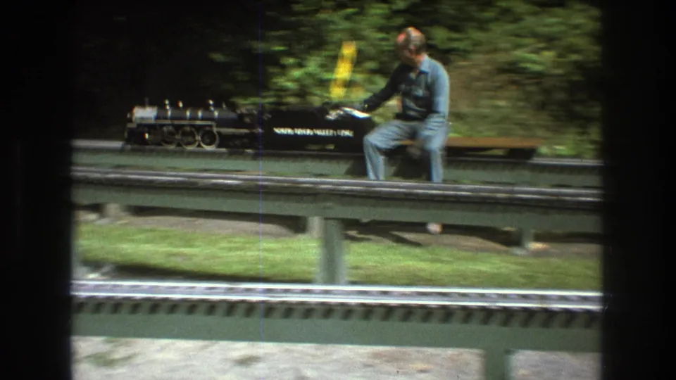 In the supplied 1975 Baxter, Maine setting, an adult man in blue clothing and a cap rides next to a black miniature steam locomotive marked with visible lettering on raised rails.