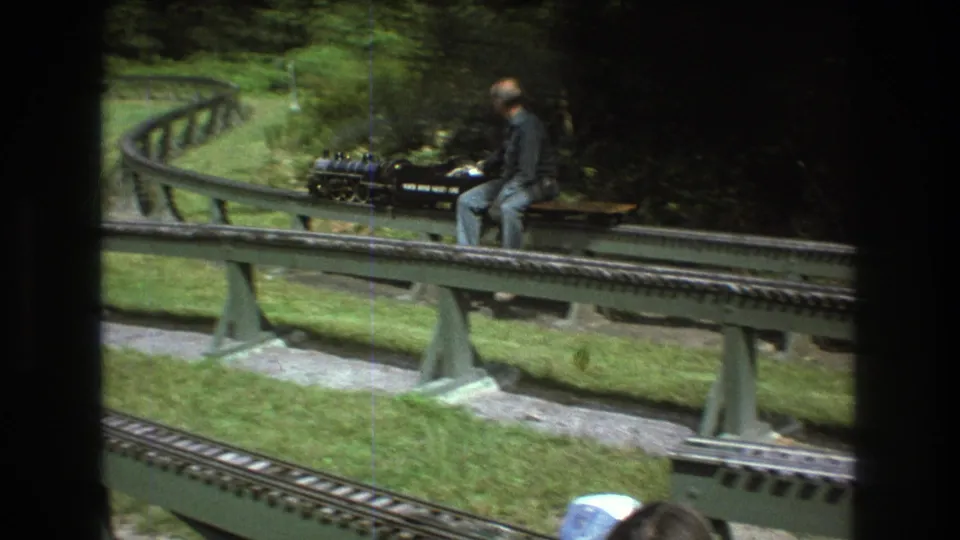 The 1975 Baxter, Maine home-movie frame shows an adult operator riding a miniature steam train around a curve, with elevated track supports, grass, and foreground rails visible.