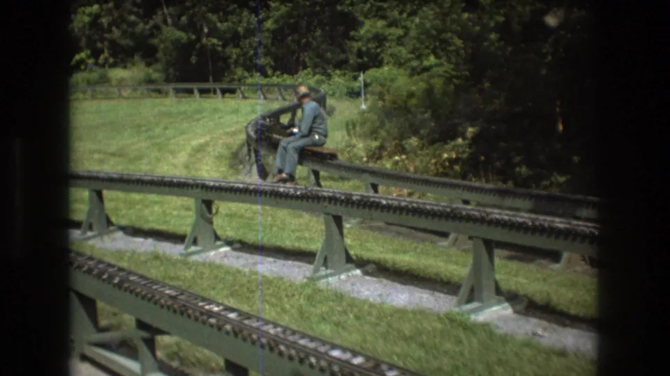 In the supplied Baxter, Maine 1975 context, the miniature train and its adult operator are seen from farther back on raised rails running through a grassy, tree-lined outdoor area.