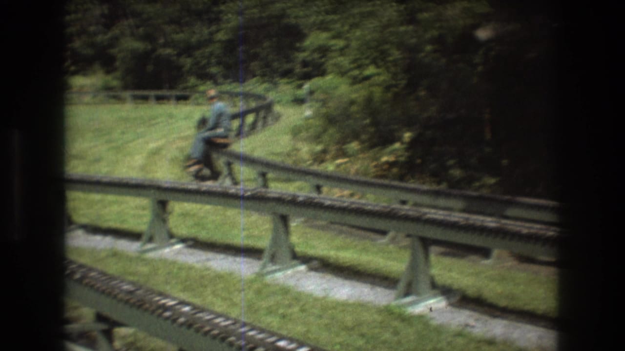 In Baxter, Maine in 1975, a rider appears seated on a miniature train car moving along raised rails through a grassy wooded park setting.