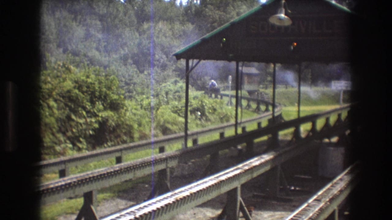 1975 8mm footage in Baxter, Maine shows a smoking train nearing the Southville station area beyond parallel tracks and a dark window frame.
