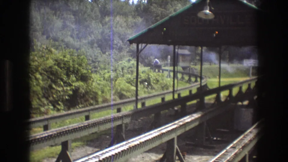 1975 8mm footage in Baxter, Maine shows a smoking train nearing the Southville station area beyond parallel tracks and a dark window frame.