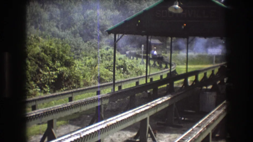 A small figure in light clothing is visible near the covered station platform as smoke hangs over the trackside greenery in the 1975 Maine rail scene.