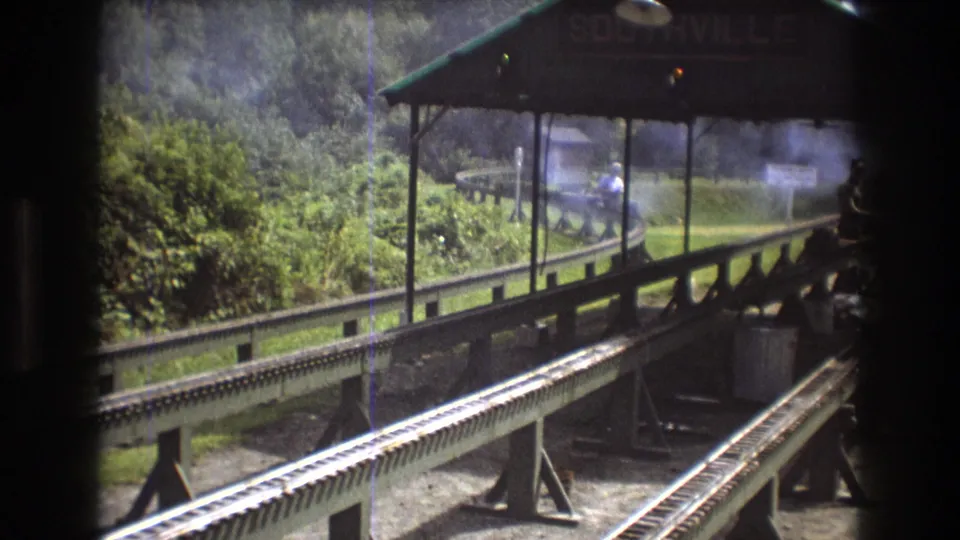 The final frame holds on the Southville station area, with parallel tracks, canopy supports, green surroundings, and smoke behind the platform in Baxter, Maine.