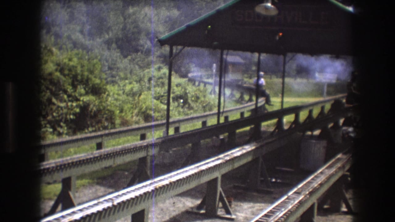 1975 Baxter, Maine 8mm frame from inside a moving train, looking past dark carriage edges toward a covered Southville platform, rail tracks, foliage, haze, and a seated adult man.
