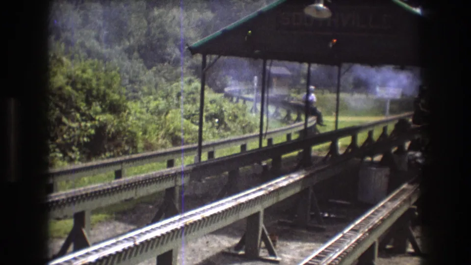1975 Baxter, Maine 8mm frame from inside a moving train, looking past dark carriage edges toward a covered Southville platform, rail tracks, foliage, haze, and a seated adult man.