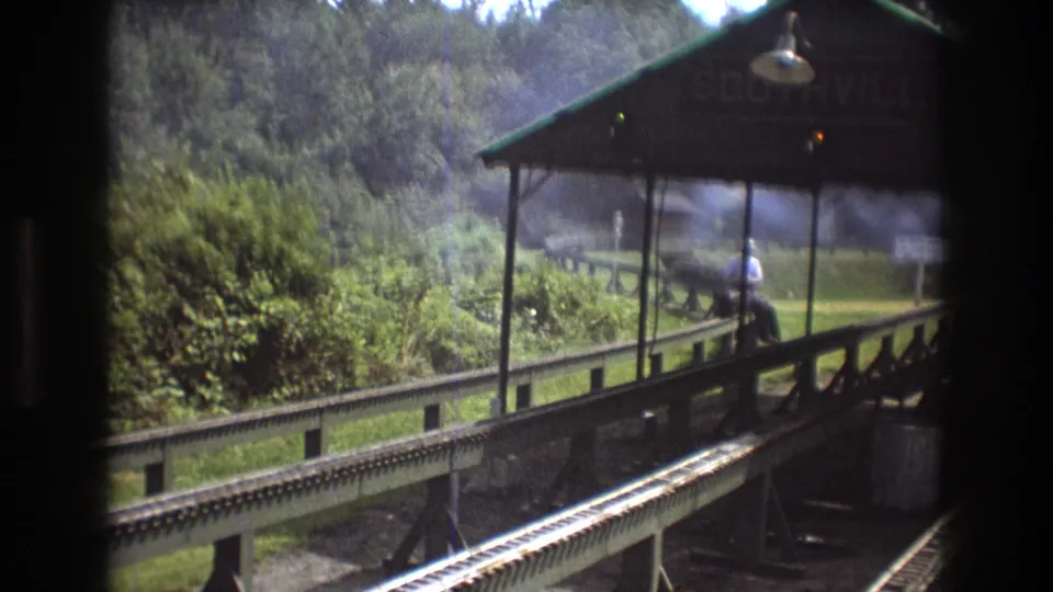 1975 Baxter, Maine frame showing the train passing a covered station platform with a faint Southville sign, an adult man by the railing, parallel tracks, and leafy wooded surroundings.
