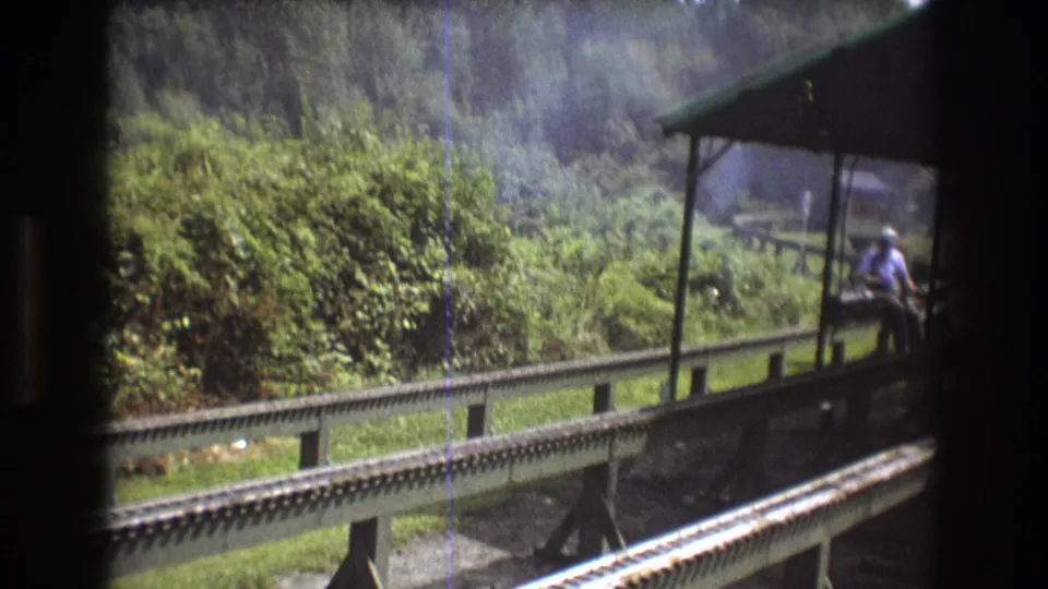 1975 Baxter, Maine 8mm still with the moving train looking across rails and a green guardrail toward dense foliage and a covered platform with an adult man partly visible at right.