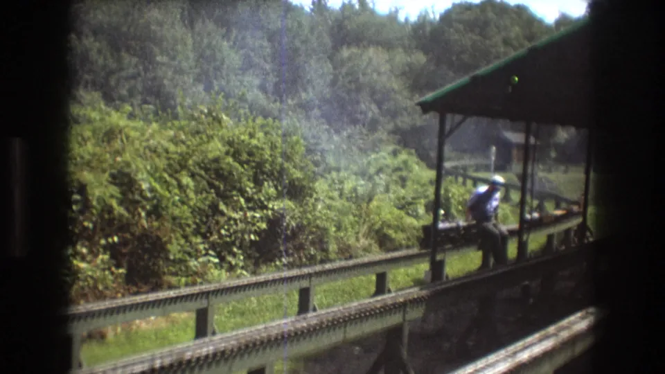 1975 Baxter, Maine frame showing an adult man seated on or near the Southville station platform while the camera passes from a moving train, with tracks, railing, trees, and film-edge shadows visible.