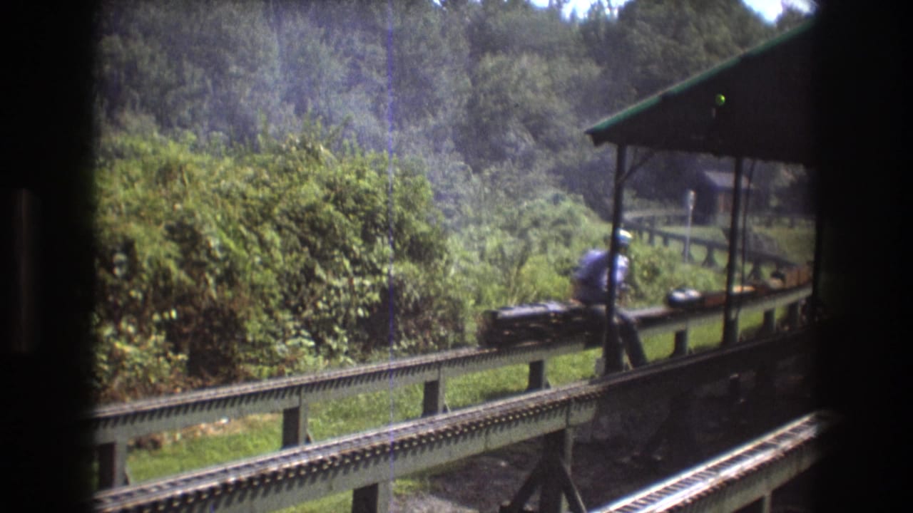 A 1975 Baxter, Maine 8mm frame shows a man in a light blue shirt and cap beside a black miniature train on elevated rails, with forest foliage behind him.