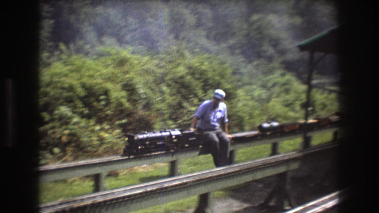 Seated operator rides beside a black miniature locomotive on an elevated track.