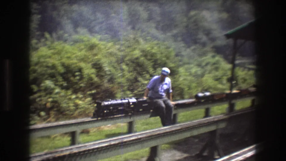 A man in a cap sits beside a black miniature steam locomotive as it travels along elevated rails through a green wooded setting in Baxter, Maine in 1975.