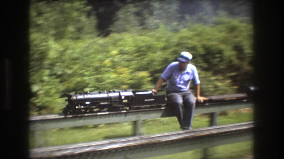 The 8mm frame shows the black miniature locomotive passing close to a seated male operator on a raised outdoor railway, with motion-softened greenery in the background.