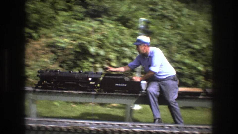 A man in a cap and light blue shirt stands beside and reaches toward a black miniature train on elevated outdoor rails, surrounded by dense green foliage in the 1975 Maine footage.