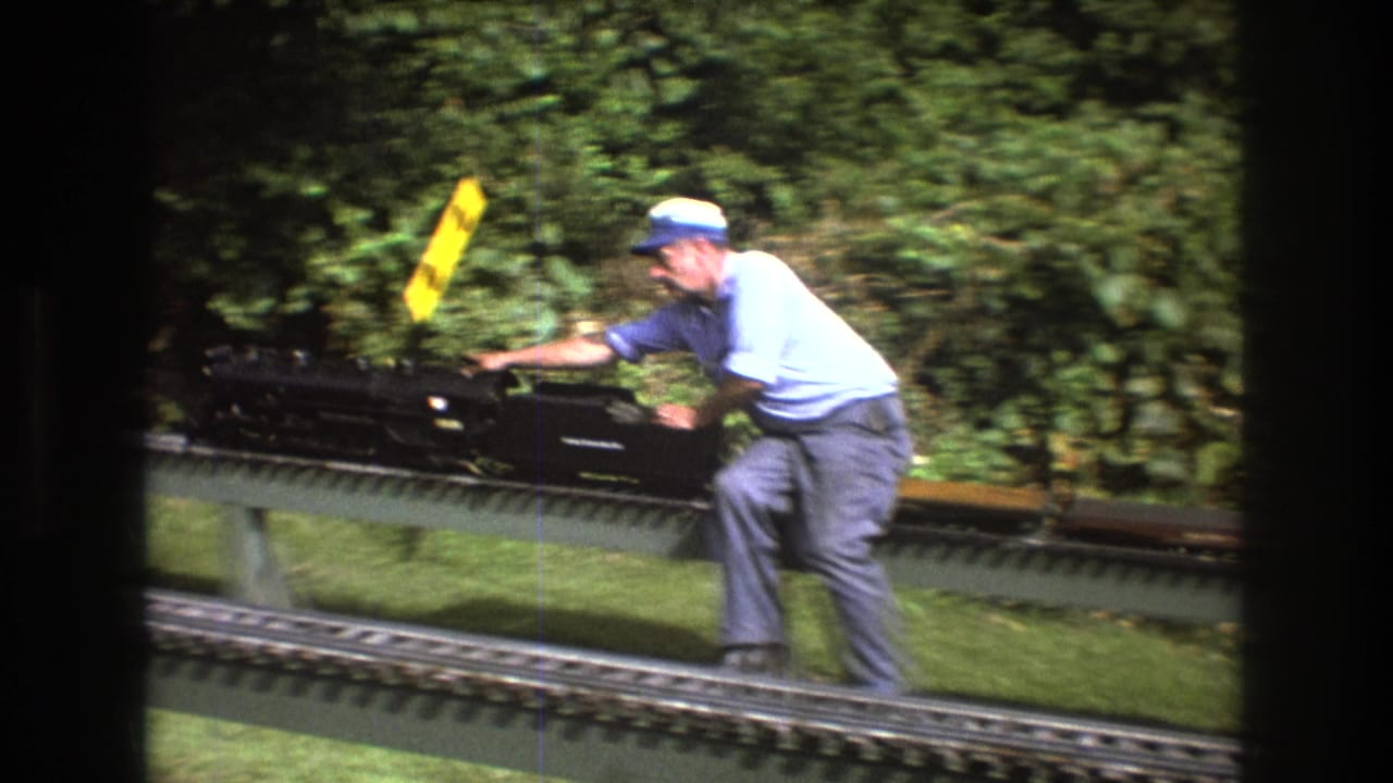 In Baxter, Maine circa 1975, an adult man in a cap leans over a black model locomotive on an elevated outdoor railway filmed through a dark window frame.