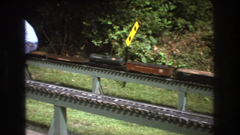 A model train with freight cars travels across elevated outdoor rails in Baxter, Maine circa 1975, with a partial view of the operator behind a window frame.