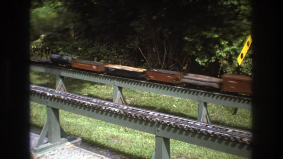Brown model freight cars roll along a raised outdoor layout in Baxter, Maine circa 1975, framed by grass, shrubs, track supports, and dark interior window edges.
