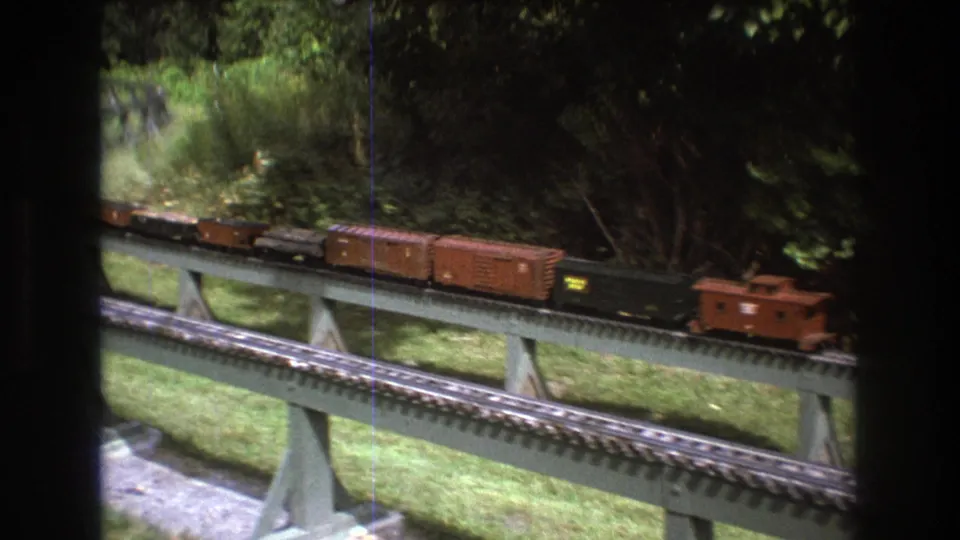 A black model locomotive, brown freight cars, and a caboose move along an elevated outdoor railway in Baxter, Maine circa 1975, seen through a window-framed home-movie view.