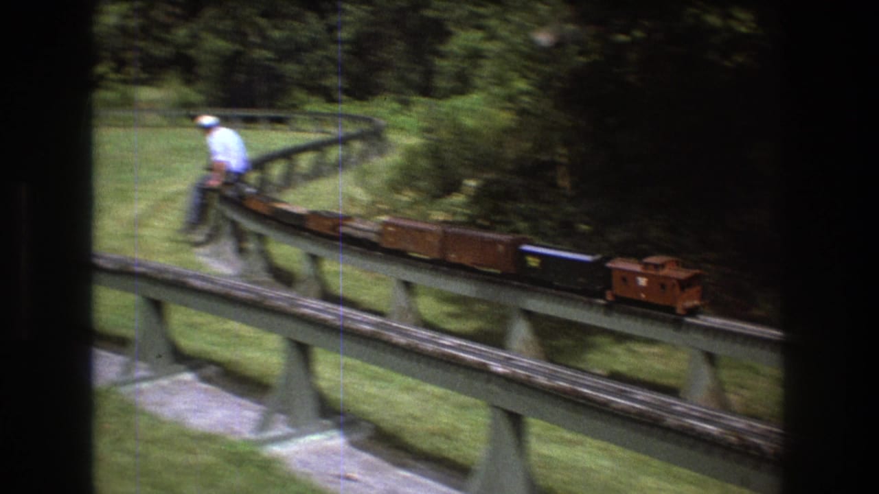 In Baxter, Maine in 1975, a man in a light shirt and white hat rides a small miniature train across an elevated track bordered by lawn and trees.