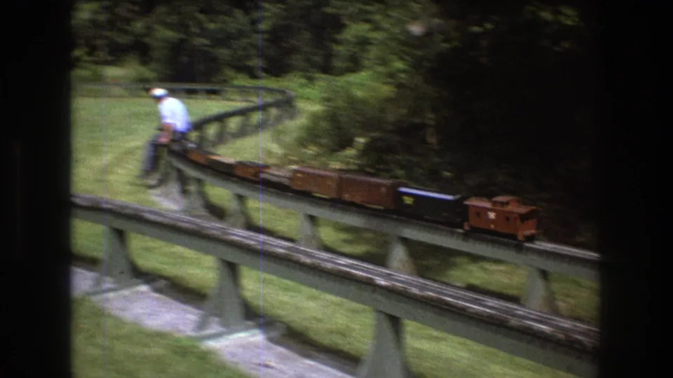 In Baxter, Maine in 1975, a man in a light shirt and white hat rides a small miniature train across an elevated track bordered by lawn and trees.