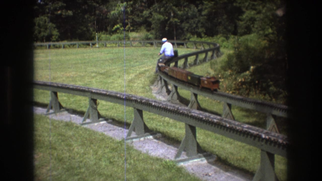 The miniature train curves along a raised outdoor railway in Baxter, Maine, with the rider seated near the front and trees enclosing the green yard.